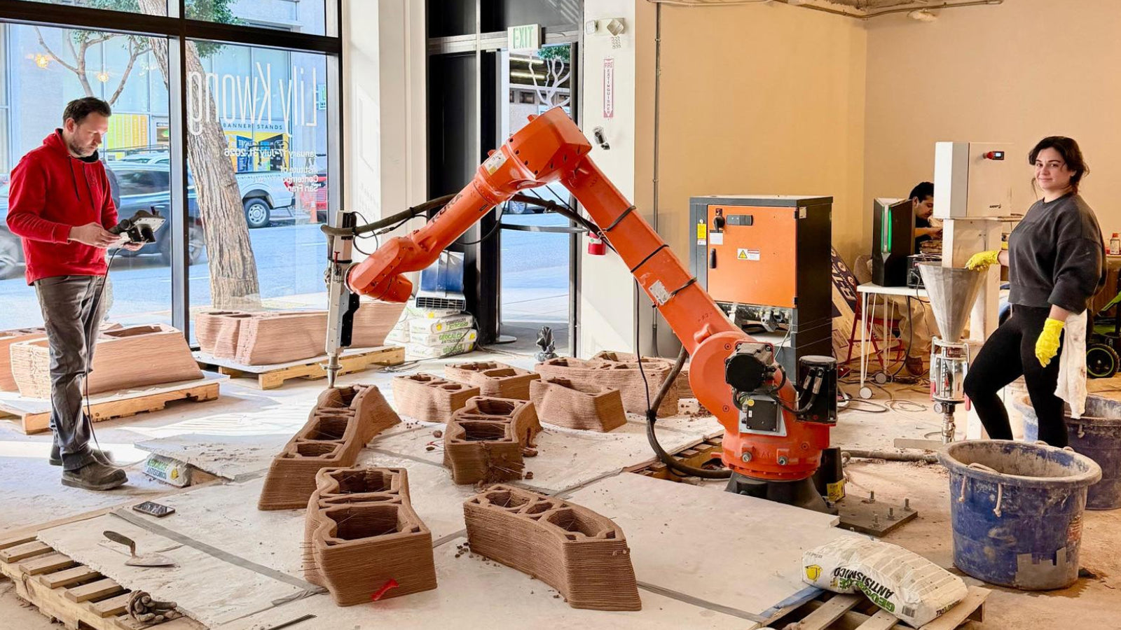 An orange robotic arm is actively working inside a workshop or studio, using a handheld tool on 3D-printed brick-like blocks. Two people are standing nearby, one in a red hoodie holding a tablet and the other in dark clothing with yellow gloves, in a space with large windows, tools, and materials.